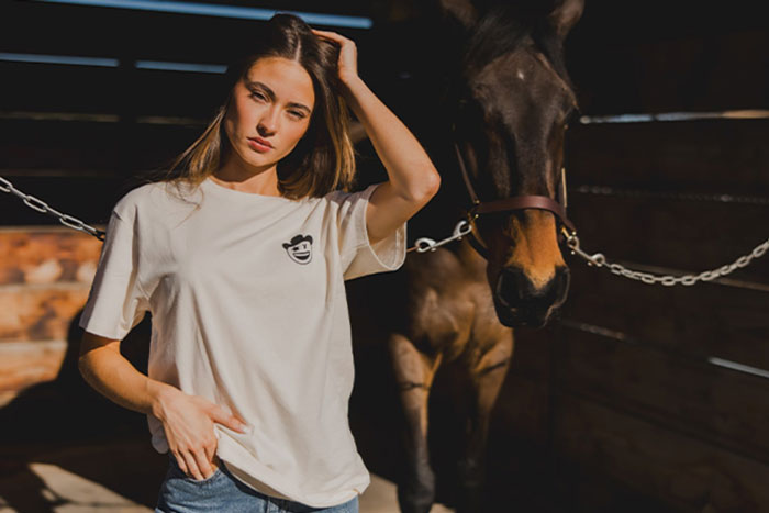 chica con camiseta de la colaboracion de milton menasco con ariat junto a un caballo castaño